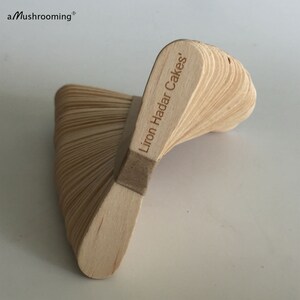 May include: A stack of light-colored wooden sticks arranged in a spiral, fan-like structure. The top stick is engraved with the text 'Liron Hadar Cakes'. The brand name 'aMushrooming' is printed in the upper left corner of the image.
