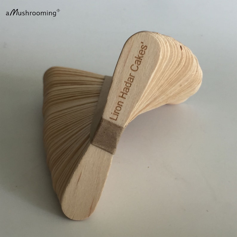 May include: A stack of light-colored wooden sticks arranged in a spiral, fan-like structure. The top stick is engraved with the text 'Liron Hadar Cakes'. The brand name 'aMushrooming' is printed in the upper left corner of the image.