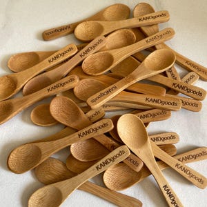 Puede incluir: Un conjunto de pequeñas cucharas de bambú de color marrón claro. Cada cuchara tiene un cuenco redondeado y un mango largo con el texto grabado "KANDgoods". Las cucharas están dispuestas sobre una superficie clara, mostrando la veta natural de la madera.