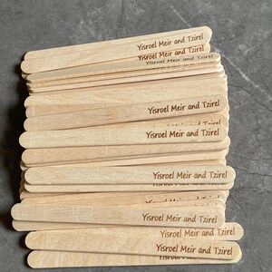 May include: A stack of light-colored wooden craft sticks, each featuring the laser-engraved text Yisroel Meir and Tzirel. The sticks are flat, rectangular, and have rounded ends, arranged in a slightly overlapping pile on a dark gray surface.