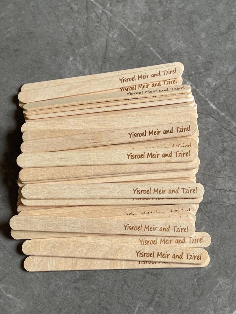 May include: A stack of light-colored wooden craft sticks, each featuring the laser-engraved text Yisroel Meir and Tzirel. The sticks are flat, rectangular, and have rounded ends, arranged in a slightly overlapping pile on a dark gray surface.