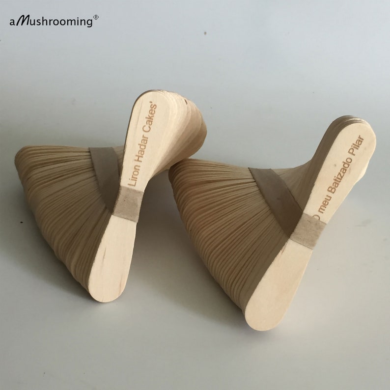 May include: Two fan-shaped structures made of stacked light-colored wooden sticks bound by a central tan band. The left structure features the text Liron Hadar Cakes, and the right structure features the text O meu Batizado Pilar. The brand aMushrooming is visible.