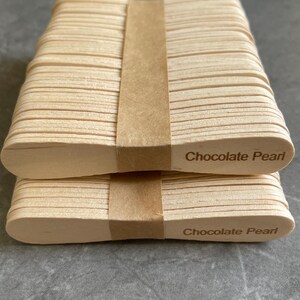 May include: Two stacks of light-colored wooden craft sticks, each bound by a brown paper band. The sticks feature rounded ends, and the text "Chocolate Pearl" is clearly printed in a dark brown font on the right side of each stack.
