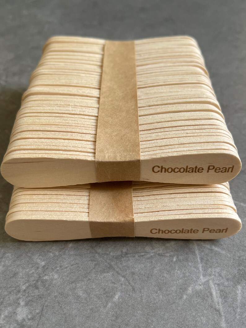 May include: Two stacks of light-colored wooden craft sticks, each bound by a brown paper band. The sticks feature rounded ends, and the text "Chocolate Pearl" is clearly printed in a dark brown font on the right side of each stack.