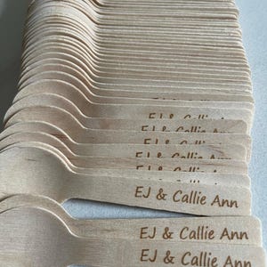 May include: A pile of wooden spoons, each with a rounded bowl and a flat handle. The handles are engraved with the text "EJ & Callie Ann". The spoons are a light brown colour and look disposable.