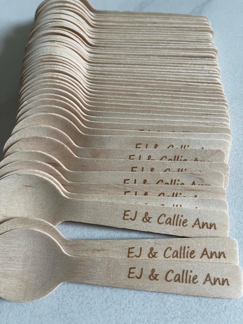 May include: A pile of wooden spoons, each with a rounded bowl and a flat handle. The handles are engraved with the text "EJ & Callie Ann". The spoons are a light brown colour and look disposable.