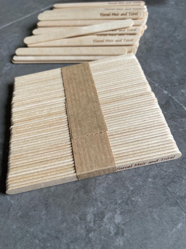May include: A stack of light-colored wooden craft sticks bundled together with a brown paper band. Each stick features the engraved text Yisroel Meir and Tzirel. The sticks are arranged in a neat rectangular pile with rounded ends.