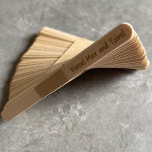 May include: A stack of light-colored wooden craft sticks arranged in a fan shape. The top stick features the engraved text Yisroel Meir and Tzirel. The sticks are smooth, flat, and have rounded ends, creating a layered, fanned-out appearance.
