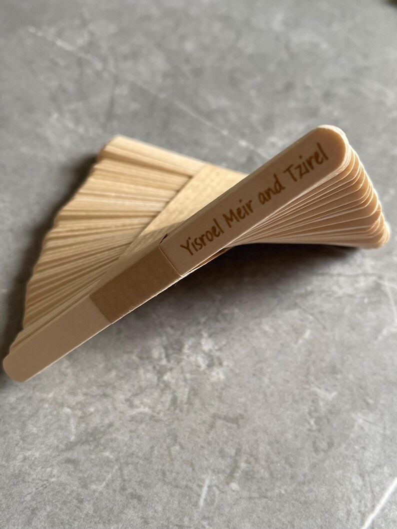 May include: A stack of light-colored wooden craft sticks arranged in a fan shape. The top stick features the engraved text Yisroel Meir and Tzirel. The sticks are smooth, flat, and have rounded ends, creating a layered, fanned-out appearance.