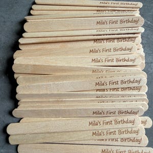 May include: A stack of light-colored wooden craft sticks, each featuring the laser-engraved text "Mila's First Birthday!" in a dark brown font. The sticks are arranged in a slightly overlapping pile, showcasing the natural wood grain texture.