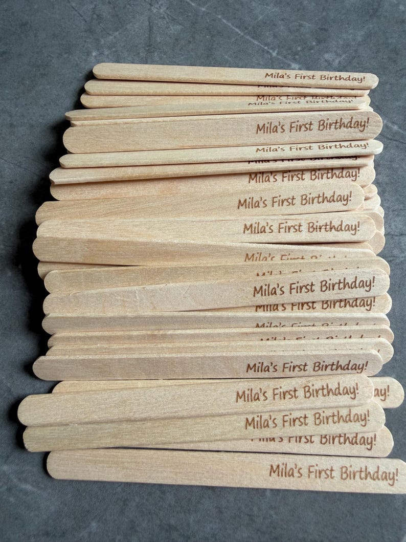 May include: A stack of light-colored wooden craft sticks, each featuring the laser-engraved text "Mila's First Birthday!" in a dark brown font. The sticks are arranged in a slightly overlapping pile, showcasing the natural wood grain texture.