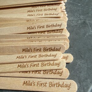 May include: A stack of light-colored wooden craft sticks, each featuring the laser-engraved text "Mila's First Birthday!" in a simple, sans-serif font. The sticks are arranged in a slightly overlapping pile, showcasing the natural wood grain texture.