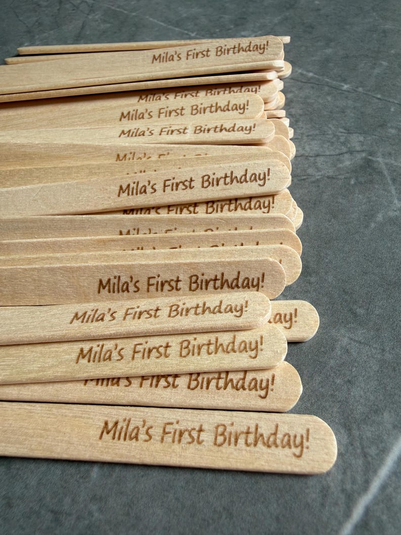 May include: A stack of light-colored wooden craft sticks, each featuring the laser-engraved text "Mila's First Birthday!" in a simple, sans-serif font. The sticks are arranged in a slightly overlapping pile, showcasing the natural wood grain texture.