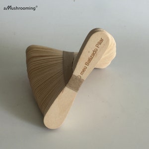 May include: A stack of light-colored wooden craft sticks held together by a tan paper band. The top stick features the laser-engraved text O meu Batizado Pilar. The brand name aMushrooming is printed in the upper left corner of the image.
