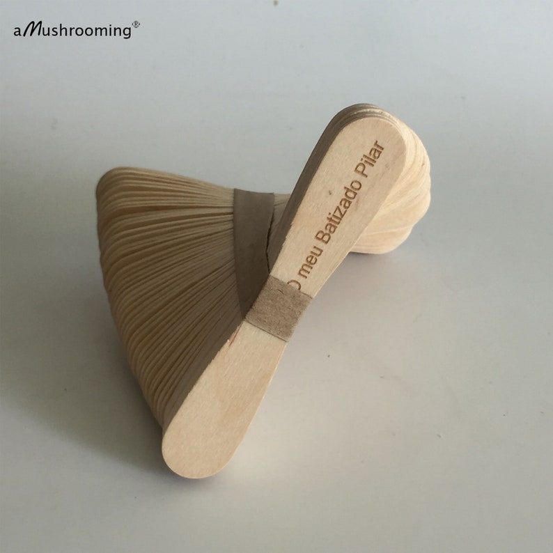 May include: A stack of light-colored wooden craft sticks held together by a tan paper band. The top stick features the laser-engraved text O meu Batizado Pilar. The brand name aMushrooming is printed in the upper left corner of the image.