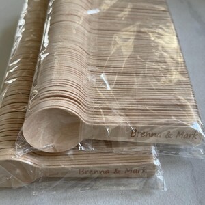 May include: Multiple packs of disposable wooden spoons, wrapped in clear plastic. The spoons are a natural wood colour and are engraved with the text "Brenna & Mark". Suitable for events or gatherings.