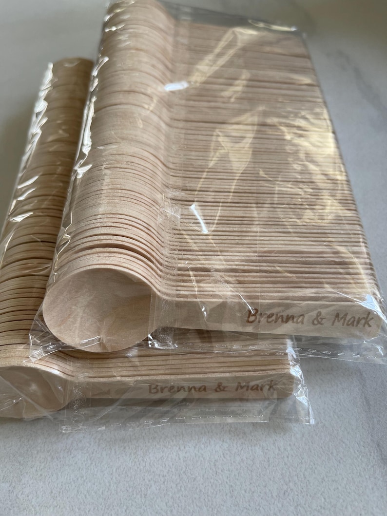 May include: Multiple packs of disposable wooden spoons, wrapped in clear plastic. The spoons are a natural wood colour and are engraved with the text "Brenna & Mark". Suitable for events or gatherings.