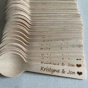May include: A pile of light-coloured wooden spoons. Each spoon is engraved with the names "Kristyna & Jon" and a heart symbol. The spoons are stacked neatly, ready for use at an event.