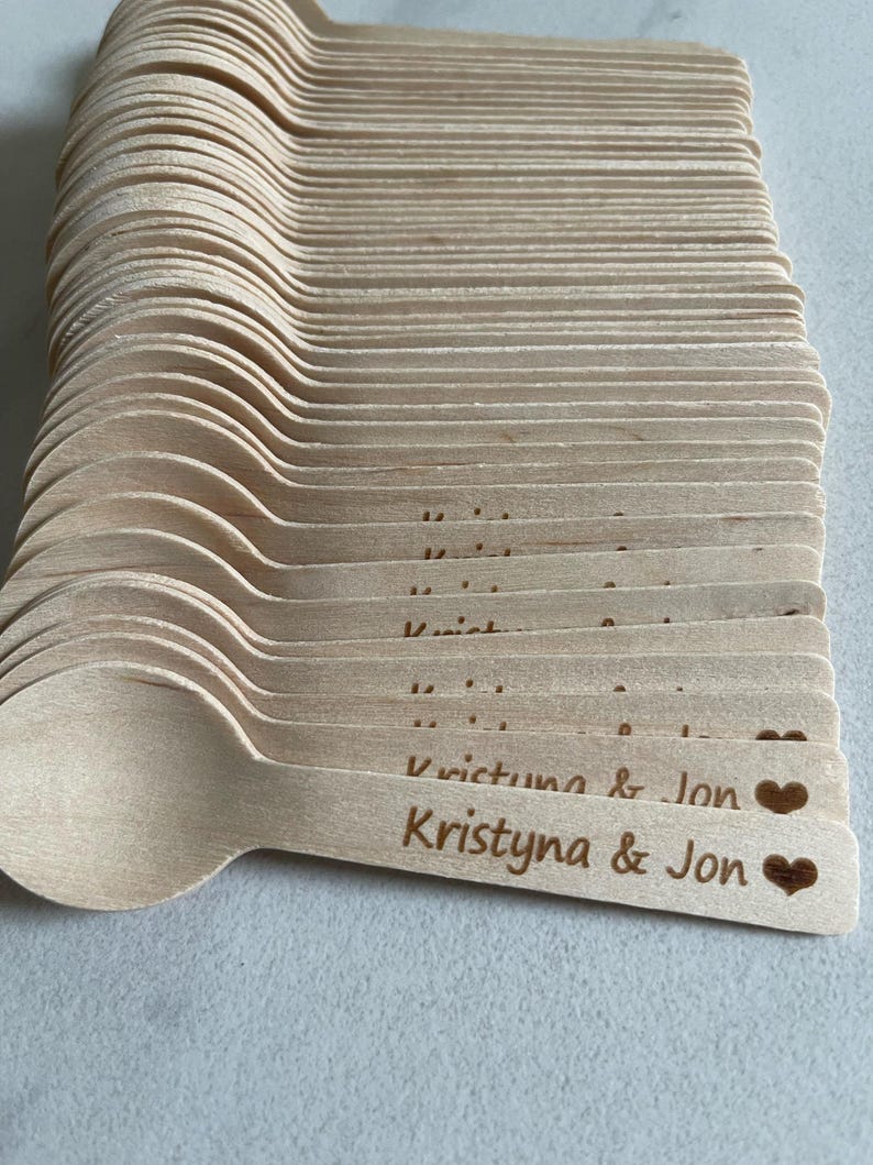May include: A pile of light-coloured wooden spoons. Each spoon is engraved with the names "Kristyna & Jon" and a heart symbol. The spoons are stacked neatly, ready for use at an event.