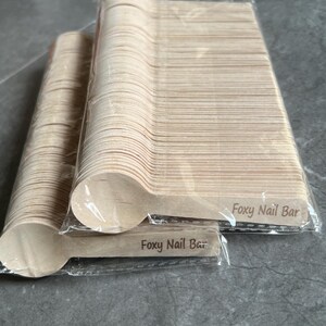 May include: A collection of disposable wooden spoons in clear plastic packaging. The spoons are light brown and have a rounded bowl shape. The text "Foxy Nail Bar" is printed on the handle of the spoons.