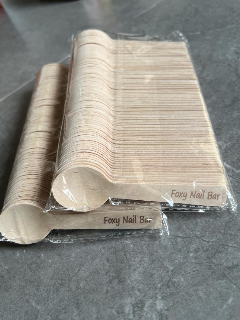 May include: A collection of disposable wooden spoons in clear plastic packaging. The spoons are light brown and have a rounded bowl shape. The text "Foxy Nail Bar" is printed on the handle of the spoons.