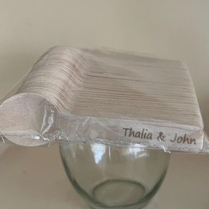 May include: A bundle of wooden spoons in a clear plastic wrapper, with the text "Thalia & John" printed on the wrapper. The spoons are light brown and are stacked above a clear glass bowl.