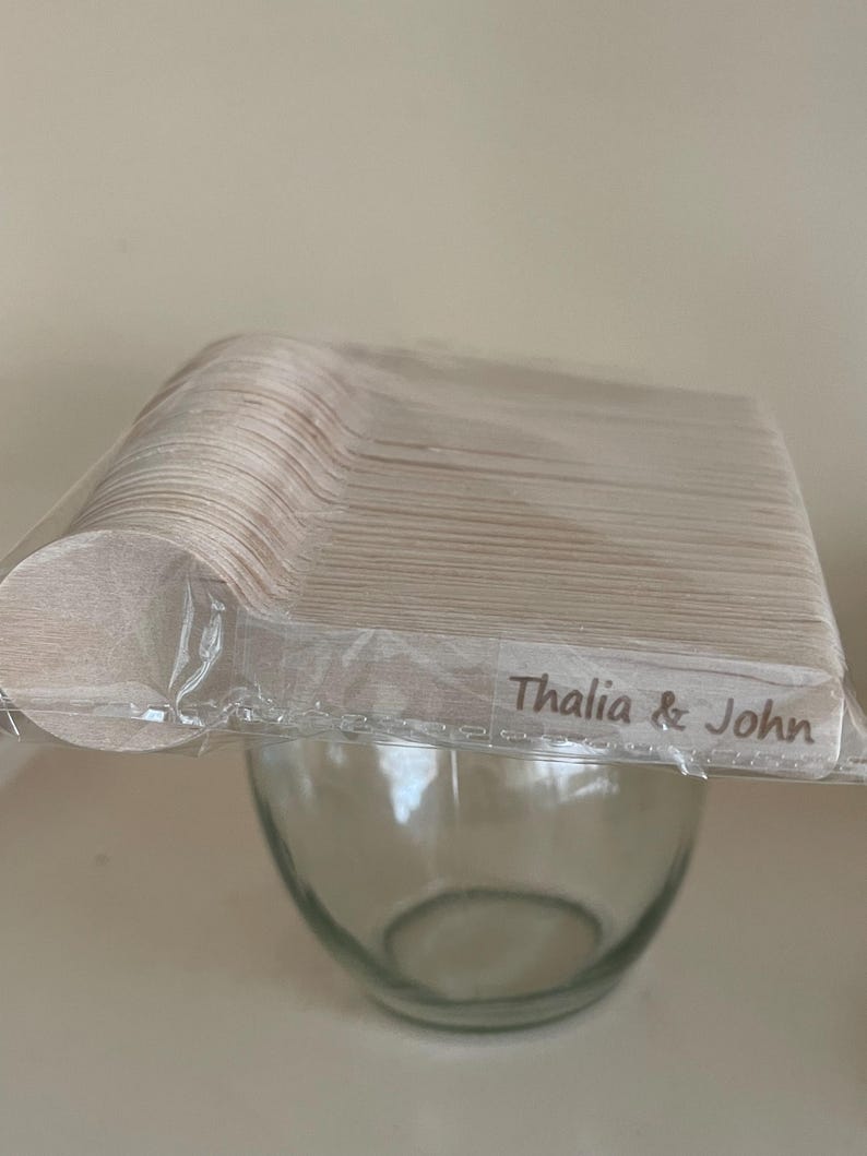 May include: A bundle of wooden spoons in a clear plastic wrapper, with the text "Thalia & John" printed on the wrapper. The spoons are light brown and are stacked above a clear glass bowl.