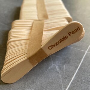 May include: A stack of light-colored wooden popsicle sticks bundled together with a brown paper band. The top stick features the text Chocolate Pearl printed in a dark brown font. The sticks have rounded ends and a smooth, natural wood finish.