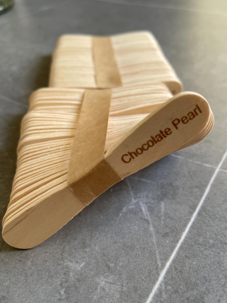 May include: A stack of light-colored wooden popsicle sticks bundled together with a brown paper band. The top stick features the text Chocolate Pearl printed in a dark brown font. The sticks have rounded ends and a smooth, natural wood finish.