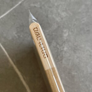 May include: A light-colored wooden stick with rounded ends, encased in a clear plastic sleeve. The wood features Hebrew text engraved vertically along the side, displaying the characters משה ורחל in a clean, sans-serif font style.