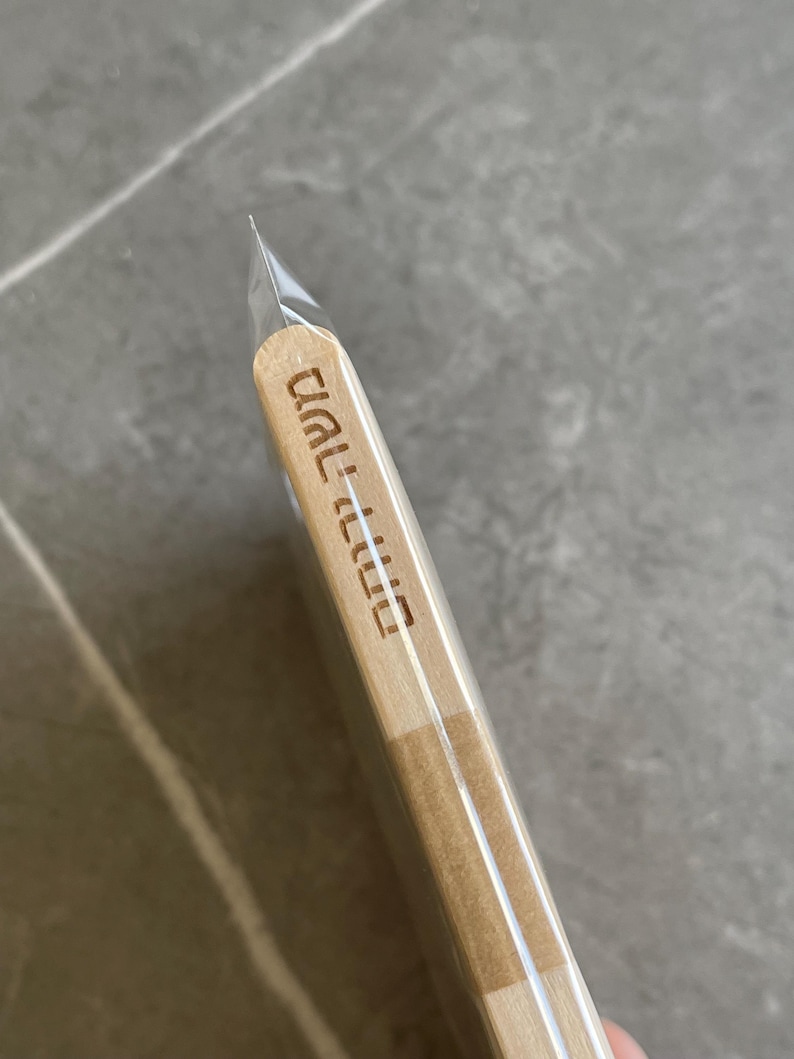 May include: A light-colored wooden stick with rounded ends, encased in a clear plastic sleeve. The wood features Hebrew text engraved vertically along the side, displaying the characters משה ורחל in a clean, sans-serif font style.