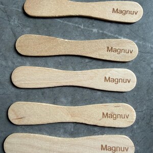 May include: Five light-colored wooden ice cream sticks arranged in a vertical stack. Each stick features a rounded, paddle-like shape and has the word "Magnuv" laser-engraved in a dark brown, sans-serif font on the right side of the surface.