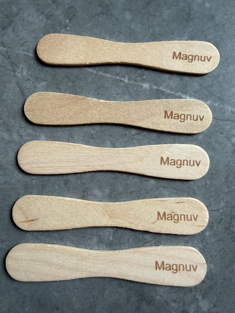 May include: Five light-colored wooden ice cream sticks arranged in a vertical stack. Each stick features a rounded, paddle-like shape and has the word "Magnuv" laser-engraved in a dark brown, sans-serif font on the right side of the surface.