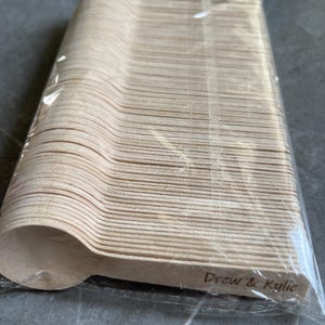 May include: A stack of disposable wooden spoons in a clear plastic wrapper. The spoons are light brown and have a simple, rounded design. The text "Drew & Kylie" is printed on the side of the spoons. Suitable for a wedding or party.