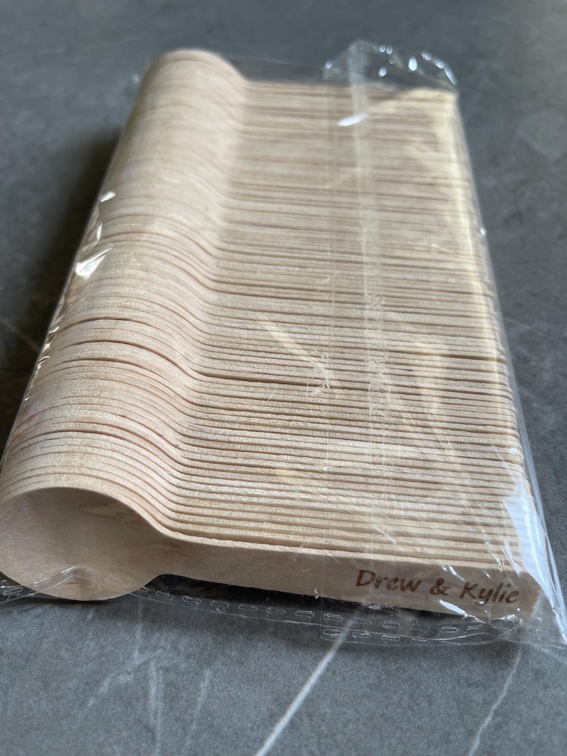 May include: A stack of disposable wooden spoons in a clear plastic wrapper. The spoons are light brown and have a simple, rounded design. The text "Drew & Kylie" is printed on the side of the spoons. Suitable for a wedding or party.