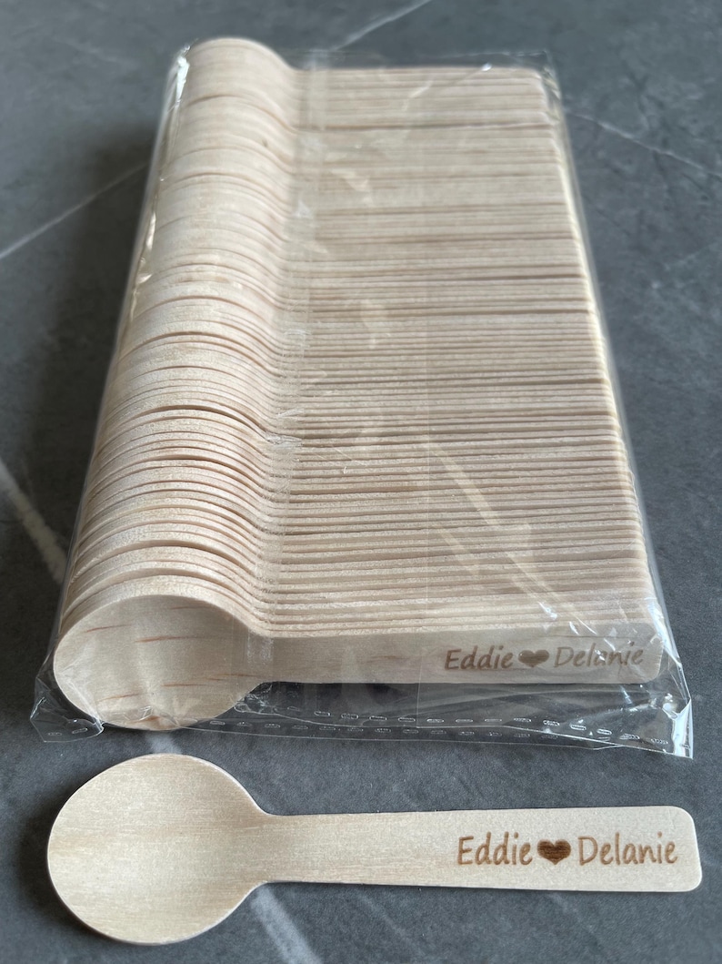May include: A bundle of wooden spoons in a clear plastic wrapper. One spoon is shown separately, with the engraved text "Eddie ❤️ Delanie". The spoons are light brown and are likely disposable.