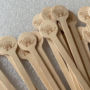 Può includere: Una collezione di bastoncini di legno per mescolare, ciascuno con una testa circolare ornata da un motivo a conchiglia e la lettera "J". I bastoncini sono di colore marrone chiaro e sembrano essere di legno.