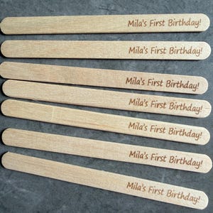 May include: Seven light-colored wooden craft sticks, each featuring the laser-engraved text "Mila's First Birthday!" in a simple, dark brown font. The sticks are arranged in a parallel, horizontal stack, showcasing a smooth, natural wood grain.