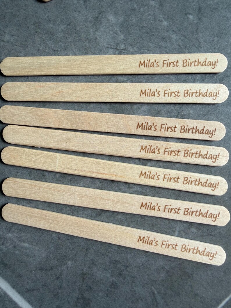 May include: Seven light-colored wooden craft sticks, each featuring the laser-engraved text "Mila's First Birthday!" in a simple, dark brown font. The sticks are arranged in a parallel, horizontal stack, showcasing a smooth, natural wood grain.