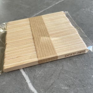 May include: A stack of light-colored wooden craft sticks bundled together with a tan paper band. The sticks are arranged in a flat, rectangular formation and are enclosed in a clear plastic sleeve. The Hebrew text "משה ירוחם" is printed on one stick.