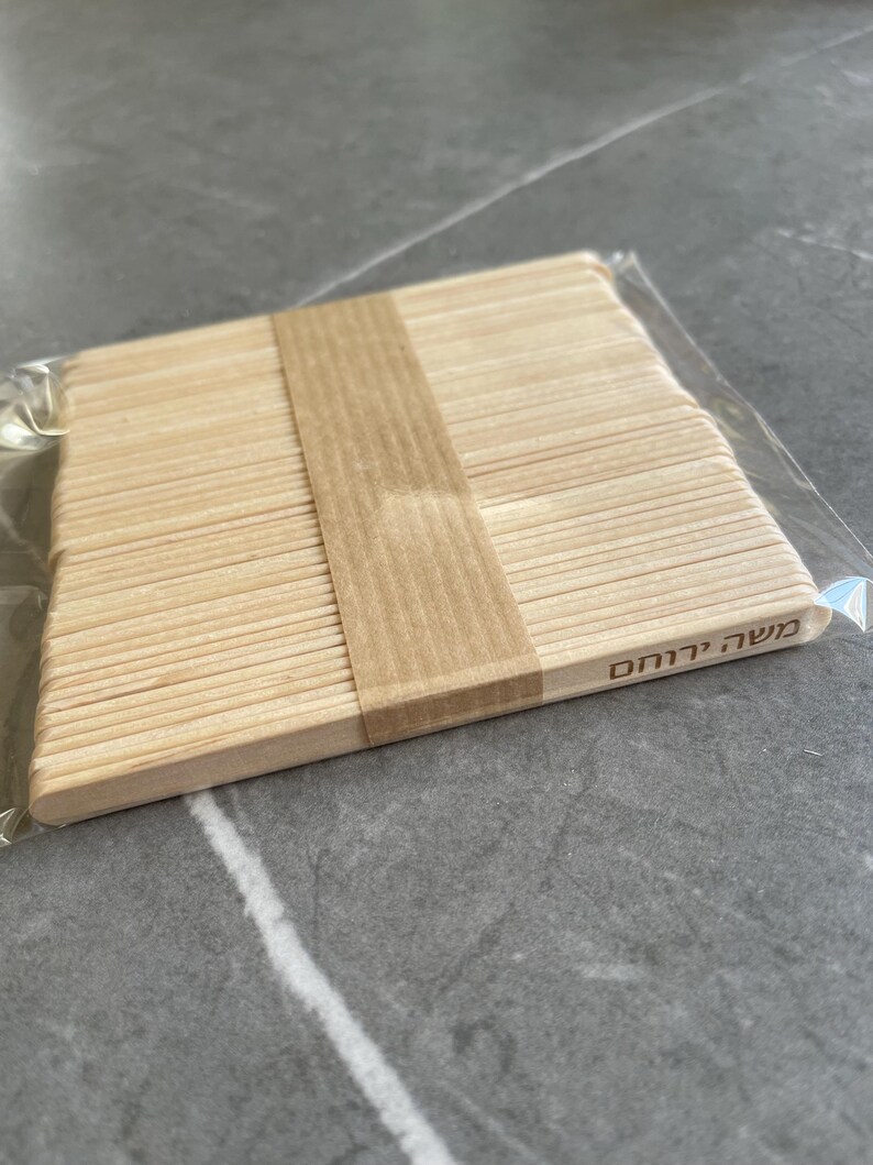May include: A stack of light-colored wooden craft sticks bundled together with a tan paper band. The sticks are arranged in a flat, rectangular formation and are enclosed in a clear plastic sleeve. The Hebrew text "משה ירוחם" is printed on one stick.