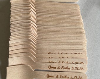 May include: A pile of light-coloured wooden spoons. Each spoon features a small, rounded bowl and a flat handle. The text "Gina & Erika 5.31.26" is engraved on the handle of each spoon. Suitable for a wedding or event.