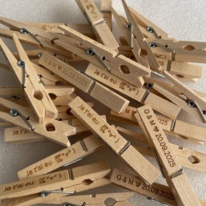 May include: A collection of wooden clothespins with metal springs. Some clothespins are engraved with the text "Je t'ai eu" and a symbol, while others have "G & M 20.09.2025" engraved on them. The clothespins are light brown.