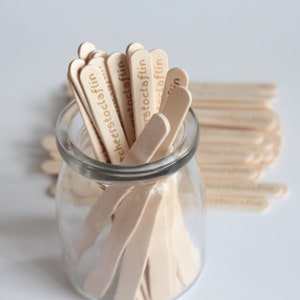 Pode incluir: Um frasco de vidro transparente cheio de palitos de madeira claros para artesanato. Cada palito apresenta o texto "cheerstoclaflin" gravado em castanho escuro. Os palitos estão dispostos verticalmente, com outros palitos deitados ao lado.