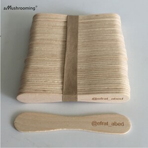 May include: A stack of light-colored wooden craft sticks secured with a brown paper band, accompanied by a single loose stick. Each stick features the text @efrat_abed printed on one end. The top left corner displays the text aMushrooming.