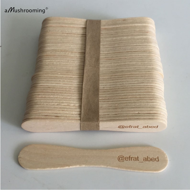 May include: A stack of light-colored wooden craft sticks secured with a brown paper band, accompanied by a single loose stick. Each stick features the text @efrat_abed printed on one end. The top left corner displays the text aMushrooming.