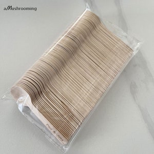 May include: A packet of disposable wooden spoons. The spoons are light brown with a natural wood grain. The text "Pazi & Jota" is printed on the handle of the spoons. The package is clear plastic, and the brand name "a Mushrooming" is in the top left corner.