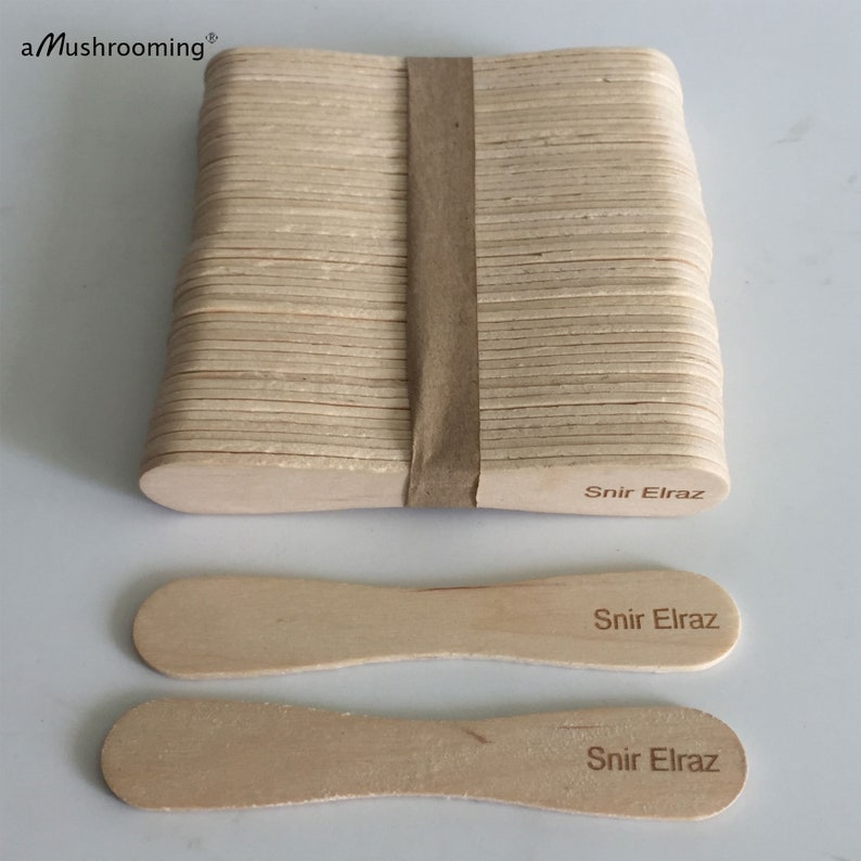 May include: A stack of light-colored wooden craft sticks secured with a brown paper band, accompanied by two loose sticks. Each stick features the laser-engraved text Snir Elraz, and the top left corner displays the text aMushrooming.