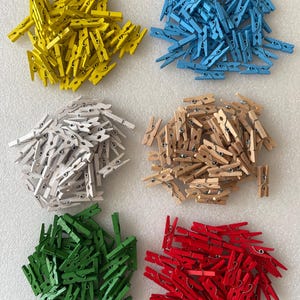 Puede incluir: Un conjunto de pequeñas pinzas de madera en seis colores diferentes: amarillo, azul, blanco, beige, verde y rojo. Las pinzas están dispuestas en montones separados, mostrando sus colores vibrantes y su diseño sencillo. Cada pinza mide unos 3 cm.