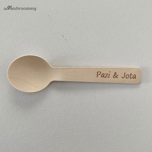 May include: A single wooden spoon with a rounded bowl and a flat handle. The text "Pazi & Jota" is engraved on the handle. The spoon is light brown in colour and appears to be made of a natural wood material. The spoon is on a grey background.
